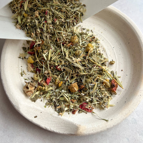 Herbal tea leaves being poured into a white bowl on a light gray background
